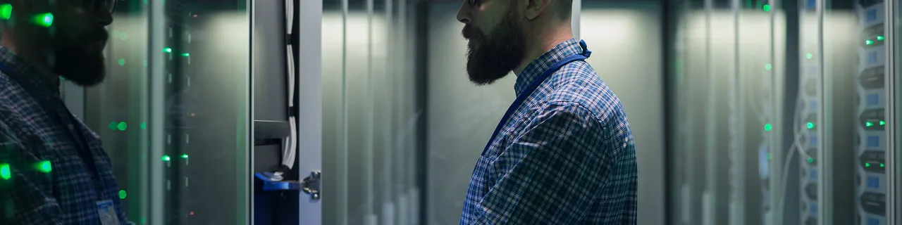 Bearded IT professional typing on laptop in a server room with glowing green racks.